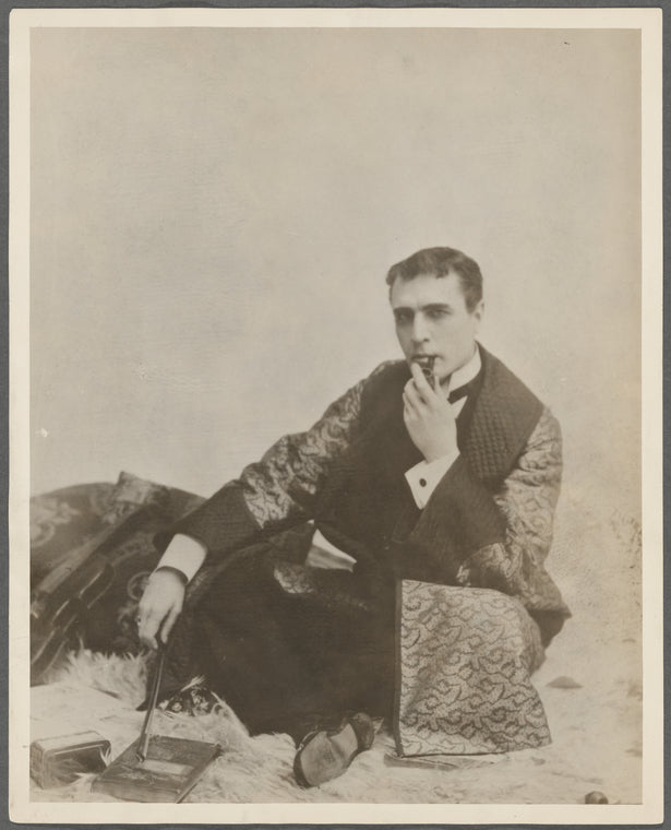 Publicity photograph of William Gillette in role of Sherlock Holmes, sitting cross-legged on fur rug, smoking a pipe