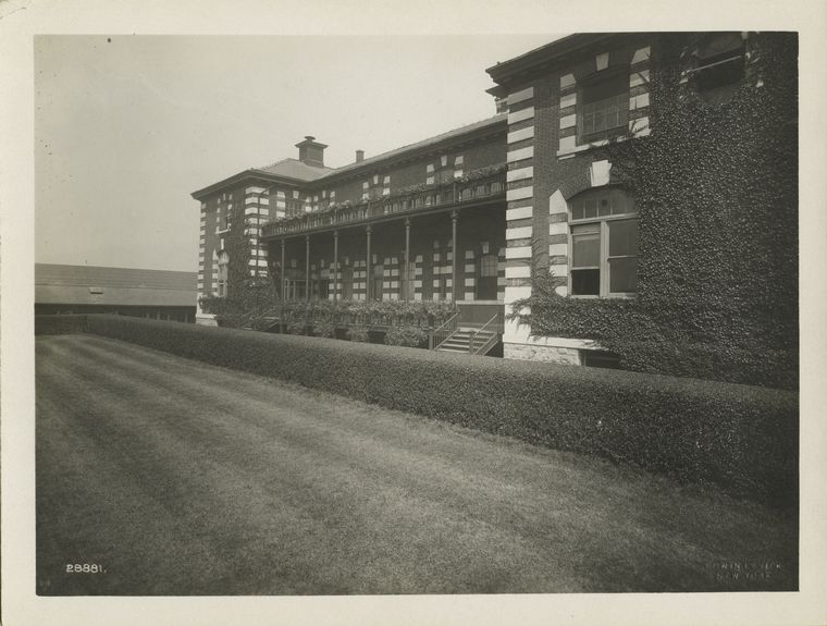 Exterior of Ellis Island building, showing ivy-covered wall, porch and balcony with flower boxes, well-kept lawn and hedge.