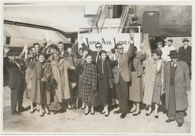 Members of Graham company in street clothes standing and waving beside a Japan Airlines plane