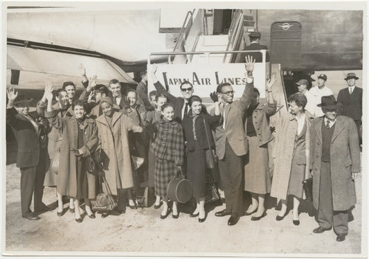 Members of Graham company in street clothes standing and waving beside a Japan Airlines plane