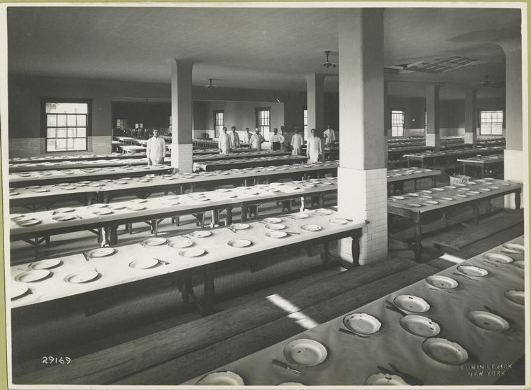 Large dining hall, empty except for about ten members of the dining hall staff. The place settings consist of a worn porcelain-enameled plate, a fork and knife.