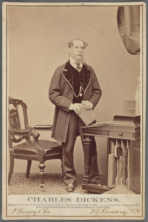 Full-length portrait of Charles Dickens, standing at desk, clasping book in both hands