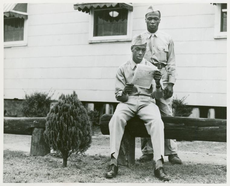 African American soldier Beverly L. Scott sitting on a wooden beam and reading a piece of paper while African American soldier Phillip S. Johnson stands behinds him and looks on, Infantry School, Fort Benning, Georgia