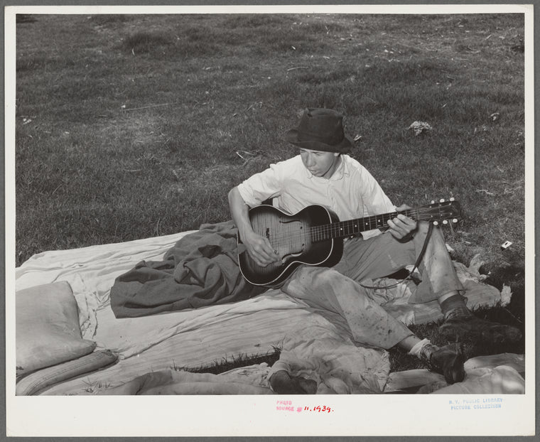 Migrant boy playing the guitar on pallet while camped near Prague, Oklahoma. Lincoln County, Oklahoma