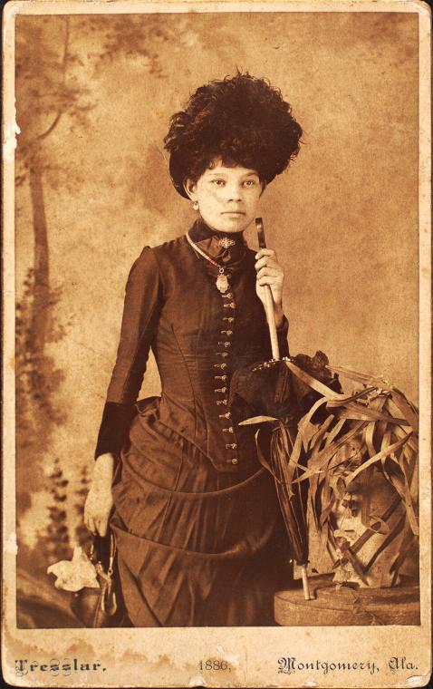 Standing woman, wearing dress with bustle, holding purse at side and umbrella resting on pedestal.