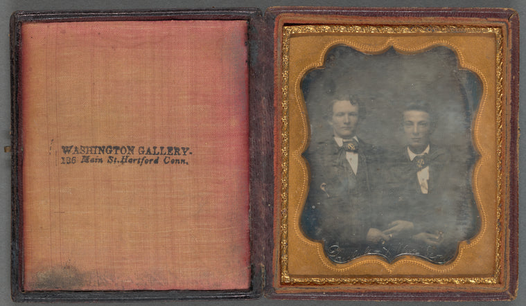 Portrait of two men seated, holding hands, wearing large bow ties, gilded with nautical symbols