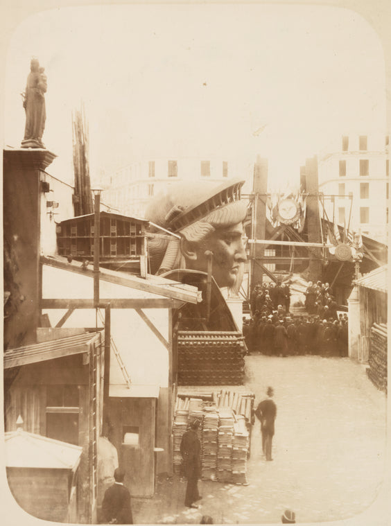 View of the external area of the workshop in Paris, showing construction materials, the head of the Statue of Liberty, and a group of men gathered in front of the left foot of the statue