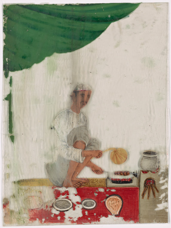 Seated male food vendor roasting meat on spit, fanning flames