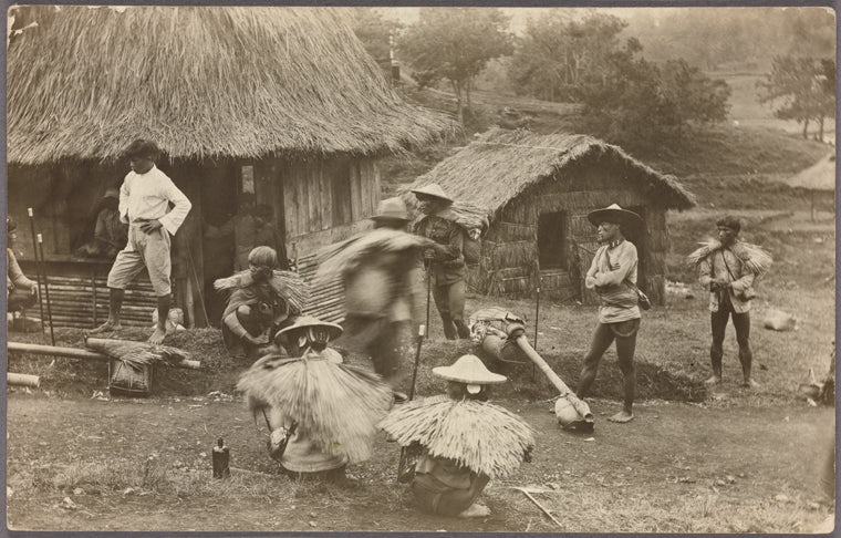 Group of individuals, some wearing thatched rain capes, in front of nipa huts.