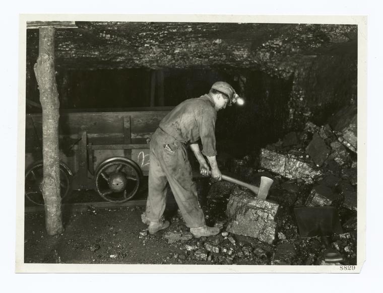 Digger is breaking large lumps of coal with an ax preparatory to loading the car. He uses an open flame lamp. Unsafe practice in gaseous mine.