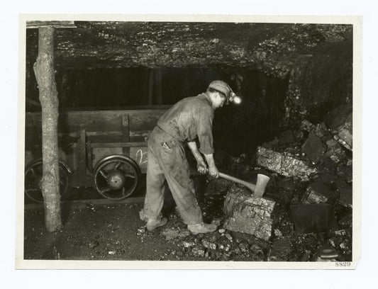 Digger is breaking large lumps of coal with an ax preparatory to loading the car. He uses an open flame lamp. Unsafe practice in gaseous mine.